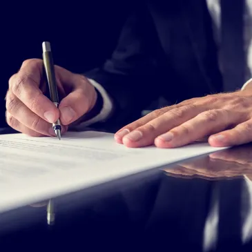 A person's hands holding a pen and signing a document on a reflective surface.